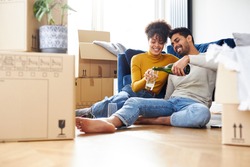 Young mixed ethnicity couple taking a break on moving day sitting on floor in lounge celebrating with champagne surrounded by removal boxes