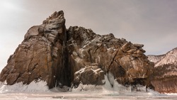 A picturesque rock without vegetation, with steep slopes and many cracks rises on a frozen lake. At the base of the cliff there are ice splashes, icicles. Sepia shades. Baikal