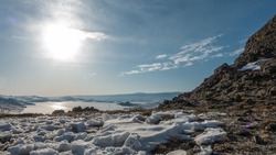 Winter landscape. A granite rock devoid of vegetation. Snow on the ground. Sun and clouds in the blue sky. In the distance, a frozen lake surrounded by mountains. Reflections on an icy surface. Baikal