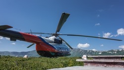 The helicopter is standing on a green meadow in the caldera of an extinct volcano. A picturesque mountain range against the blue sky. In the foreground are the steps of a wooden staircase. Kamchatka