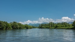 A peaceful blue river flows between the banks with lush green vegetation. In the distance, against the background of the azure sky and clouds, a mountain range is visible. Kamchatka