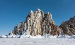 A picturesque granite rock, devoid of vegetation, rise above a frozen lake. There are cracks on the stones. There is a layer of icicles at the base. Clear blue sky. Baikal 
