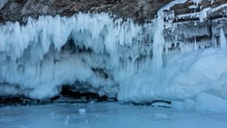 Rows of stalactite icicles, similar to lace frills, hang from the base of the granite rock. A low dark cave goes deep. Fragments of ice on the surface of a frozen lake Close-up. Baikal