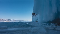 The base of the rock is covered with thick layers of splashes and icicles. Blocks of ice on a frozen lake. In the distance, against the blue sky, a mountain range. Baikal