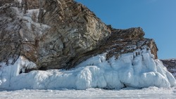 A picturesque granite rock, devoid of vegetation, rises above a frozen lake.  Cracks are visible on the stones, the texture of the stone. The base is covered with a layer of fancy icicles. Baikal