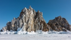 A picturesque granite rock, devoid of vegetation, rises above a frozen lake against a blue sky. Cracks on the stones.  The base is covered with a layer of icicles. Shamanka Mountain. Baikal