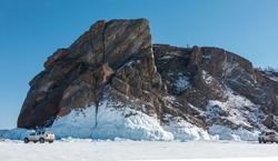 Granite rock, devoid of vegetation, against a blue sky. There is snow on the steep slopes, a thick layer of icicles at the base. There are cars on the ice of a frozen lake. Baikal