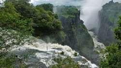 The Zambezi River flows into the gorge, bubbling and foaming. The rapid flow of water between the steep slopes. Fog over the abyss. Lush green vegetation around. Victoria Falls. Zimbabwe.             