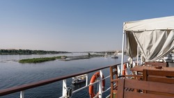 Wooden tables and chairs are installed on the upper deck of the cruise ship. From the deck you can see the Blue Nile River, green shores and the island.Boats on the water. Clear azure sky. Egypt