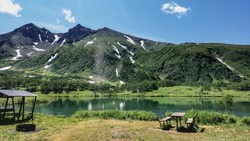 On the shore of a calm alpine lake there are places to relax, canopies, benches. A mountain range against the blue sky. Reflection in the water. Kamchatka. Vachkazhets. Lake Tahkoloch