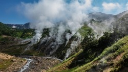 Columns of steam from hot springs rise above the slopes of the mountain. The stream flows in a hollow along a rocky bed. Blue sky. Kamchatka. Valley of Geysers