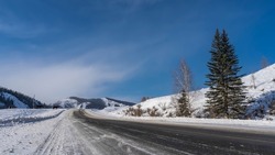 The highway goes forward through a snow-covered valley. Trees on the side of the road. The silhouette of a horse is visible by the road. In the distance, against the blue sky - a mountain. Altai.