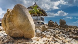 A gazebo with a thatched roof stands on a rocky hill. Wooden chairs are visible behind the latticed railing. The palm tree is nearby. Clouds in the blue sky. Huge boulders in the foreground.Seychelles