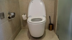 A white toilet bowl in the restroom. The lid is lifted. The walls and floor are lined with beige tiles. Rolls of toilet paper, a wash brush are visible.