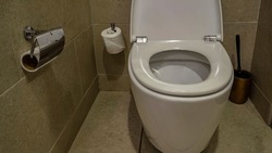 White ceramic toilet bowl with the lid raised. The walls and floor of the  restroom are lined with beige tiles. Rolls of toilet paper, a washing brush are visible.