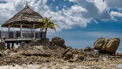 Granite boulders of the seabed were exposed at low tide. The gazebo with a thatched roof is surrounded by a lattice fence. The palm tree is nearby. Clouds in the blue sky. Seychelles. Mahe