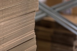 Detail of edge of neatly stacked corrugated cardboard for making boxes, with metal factory shelves in background in soft-focus