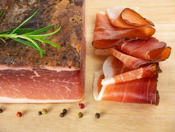 Speck with slices on the cutting board. Typical South Tyrolean smoked bacon. Sliced raw ham. Dry cured meat. Traditional cold cuts, Italian speck with rosemary and pepper.