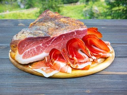 Whole speck with slices. Typical South Tyrolean raw ham outdoors. Italian food. Table in nature, in the woods.