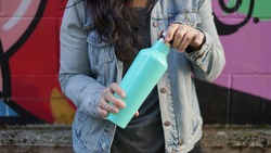Woman opening a stainless steel water bottle. Her head is out of frame, she's wearing a jean jacket and behind her is a graffiti wall.                             