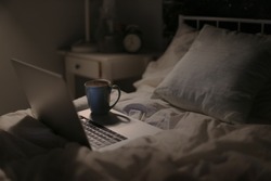 working at night in bed online from home with coffee on the laptop and background of bedroom with calm light during quarantine and corona virus crisis
