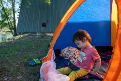 A child sits in a tent on the lakeshore, watches cartoons on the phone and eats sweets, a girl sits in a tent, tourism, family, summer, vacation, picnic, travel