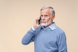 Serious senior businessman calls to employee on phone standing on beige background. Elderly man with grey beard resolves work issues via smartphone