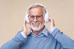 Studio portrait of relaxed cheerful senior man in glasses wearing  holding wireless headphones isolated on beige wall, enjoying listening to favourite music soundtrack, wellbeing, meditation concept