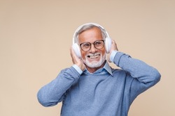 Studio portrait of relaxed cheerful senior man wearing and holding wireless headphones isolated on beige wall, enjoying listening to favourite music soundtrack, wellbeing, meditation concept