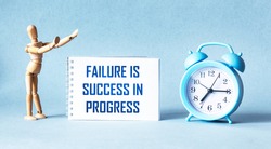 Failure is success in progress - inspirational text on a notebook, next to a wooden doll clock on a blue background.