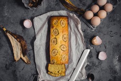 Homemade sliced banana bread in baking tray ready to serve on breakfast, egg shell walnuts, linen seeds and knife, spoon , whisk on dark grey concrete background. Top view