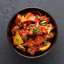 Chicken Manchurian in bowl at black concrete background. Chicken Manchurian is Indian Chinese cuisine dish with Chicken breasts, bell pepper, tomatoes, soy sauce.