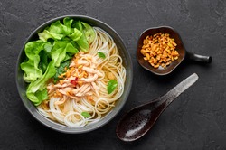 Guay Tiew Gai Cheek or Thai Chicken Noodle Soup in black bowl on dark slate backdrop. Guay Tiew Gai is Thailand cuisine soup with rice noodles, chicken meat, sauces, greens. Thai food. Top view