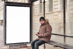 Portrait of young man in casual winter clothes with glasses and mobile phone on stop bus. An attractive teenager sits in old town waiting for public transport on frosty day. Copy space for site