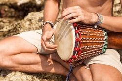 Close up hands young man drummer, playing rhythm with djembe drum, sitting at tropical coral reef coast background. Travel vacation and music relaxation concept. Selective focus. Copy text space
