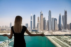 Rear view of young cute woman in black on balcony with view of skyscrapers Dubai UAE, pensive looking. Lovely lady posing from behind on terrace of tower block. Leisure activity concept. Copy ad space