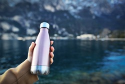 Close-up of female hand holding reusable, steel thermo shiny bottle for water, on the background of clear water of a lake with a turquoise hue. Copy space concept. 
