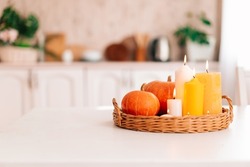 Yellow pumpkins and beautiful yellow and white candles in a wicker tray on a white table in the interior of a home kitchen. Cozy autumn concept