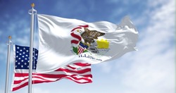 The Illinois state flag waving along with the national flag of the United States of America. In the background there is a clear sky. Illinois is a state in the Midwestern region of the United States