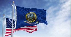 The Idaho state flag waving along with the national flag of the United States of America. In the background there is a clear sky. Idaho is a state in the Pacific Northwest region of the United States