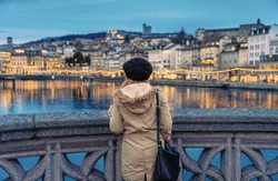Blonde girl with wool hat and heavy clothing staring at the Limmat river in central Zurich at dusk in winter