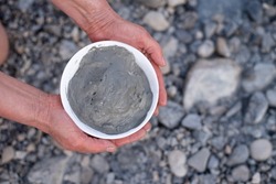 Woman's hands holds bowl with gray medical mud on rocky shore background. Healthy vacation on Dead Sea with therapeutic mud on face and body. Travel to hydrogen sulfur hot water in Israel