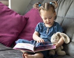 Child reading. Little cute girl 1-3 is reading a book with a toy plush hare sitting on a sofa