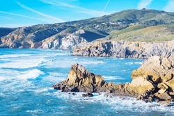 Pictorial view of blue lagoon, rocky beach seashore coastline, foamy sea waves surrounded by mountains cliffs on windy sunny day. Summer, holiday, vacation, travelling, seascape, tourism, shoreline.
