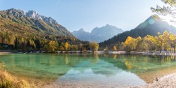Panorama of a mirror reflection in Lake Jasna, surrounded by colored trees in the fall, and Triglav mountains in the background