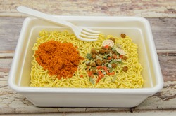 Instant dryed noodles with dry vegetables, spices with red pepper in disposable container with plastic fork. Traditional cheap Asian food. Fast food, raw noodles. Selective focus, blurred background