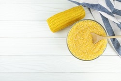 Corn grits in a glass bowl with a wooden spoon, a cob of boiled corn and a tea towel on light wooden boards. Corn products. Background for adding text.