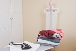 Housewife's workstation for ironing linen. Ironing board with iron, vertical household garment steamer and a stack of clean linen. Preparing for ironing.