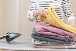 Woman stands by an ironing board with an iron and is folding a stack of ironed, clean terry towels. Housework, ironing of the washed linen. The housekeeper is engaged in household chores.