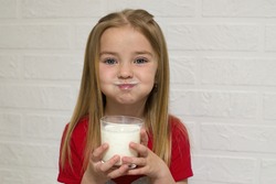 The baby shows a milk moustache and big cheeks. the girl holds a glass of milk, natural yogurt in her hand